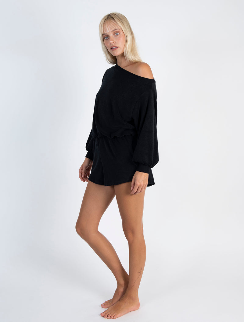 LVHR Malia Off the Shoulder Romper in black. Organic recycled terry cloth knit with rib cuffs and neckline, front pockets and elastic waist. Off the shoulder styling. Side.